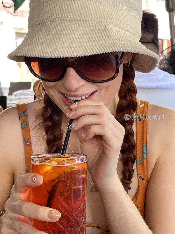 全画幅图片，迷人的红头发的女人喝着Spritz Veneziano鸡尾酒饮料与塑料吸管，冰块和橙子片，户外戴着太阳镜，遮阳帽和太阳裙与穆拉诺玻璃手镯，户外用餐设置背景